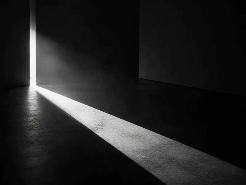 A thin blade of hard white light through a narrow slot in a black wall, cutting a sharp wedge across a dark floor.